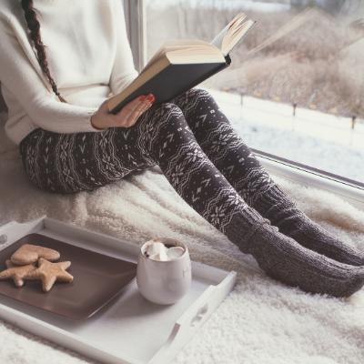 Girl reading on a windowsill with hot cocoa and cookies on a tray.